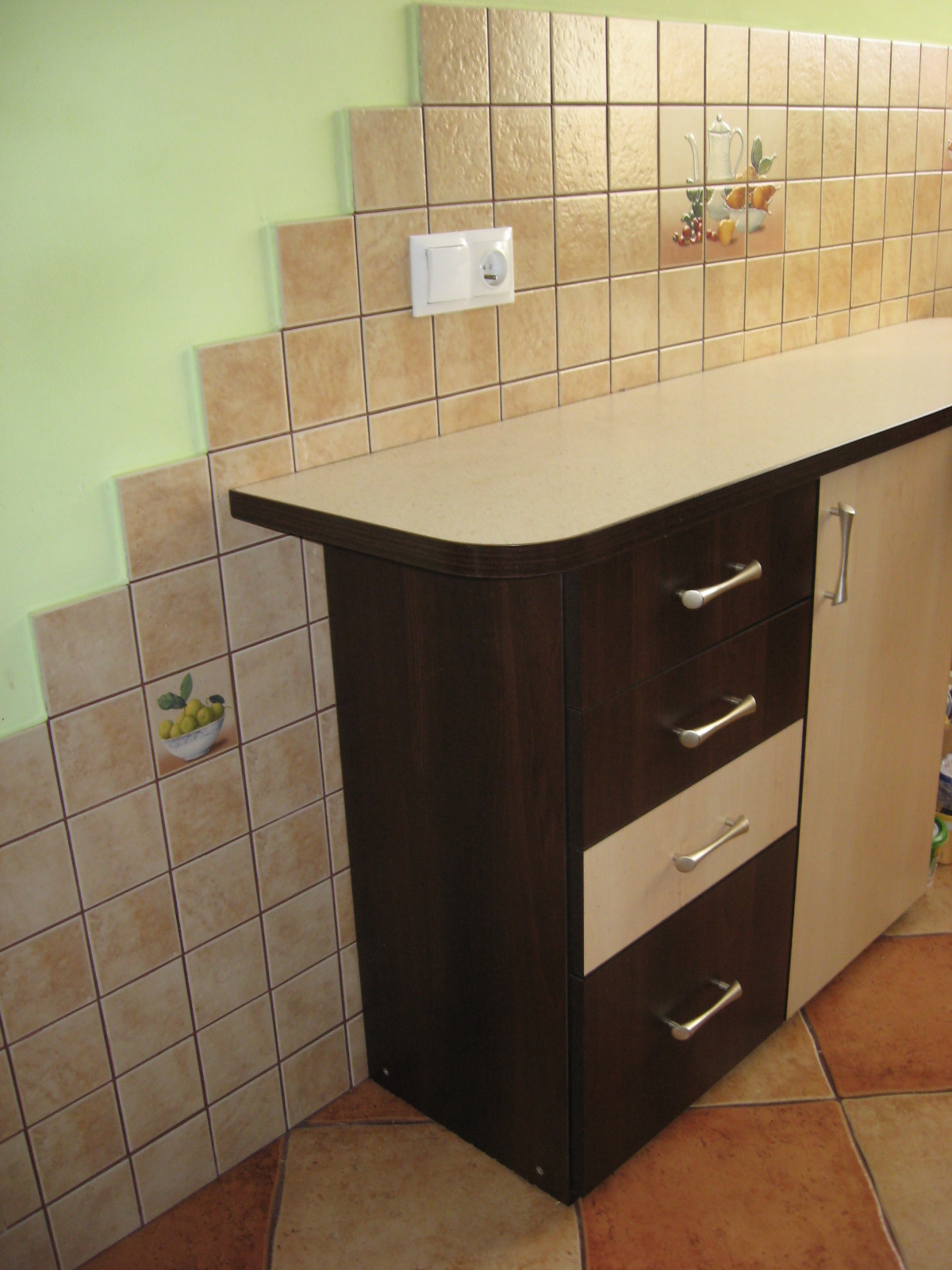 Fragment kuchni z beżowymi płytkami z motywami owocowymi, brązowy słupek z szufladami i jasnym blatem, widoczne gniazdko elektryczne.